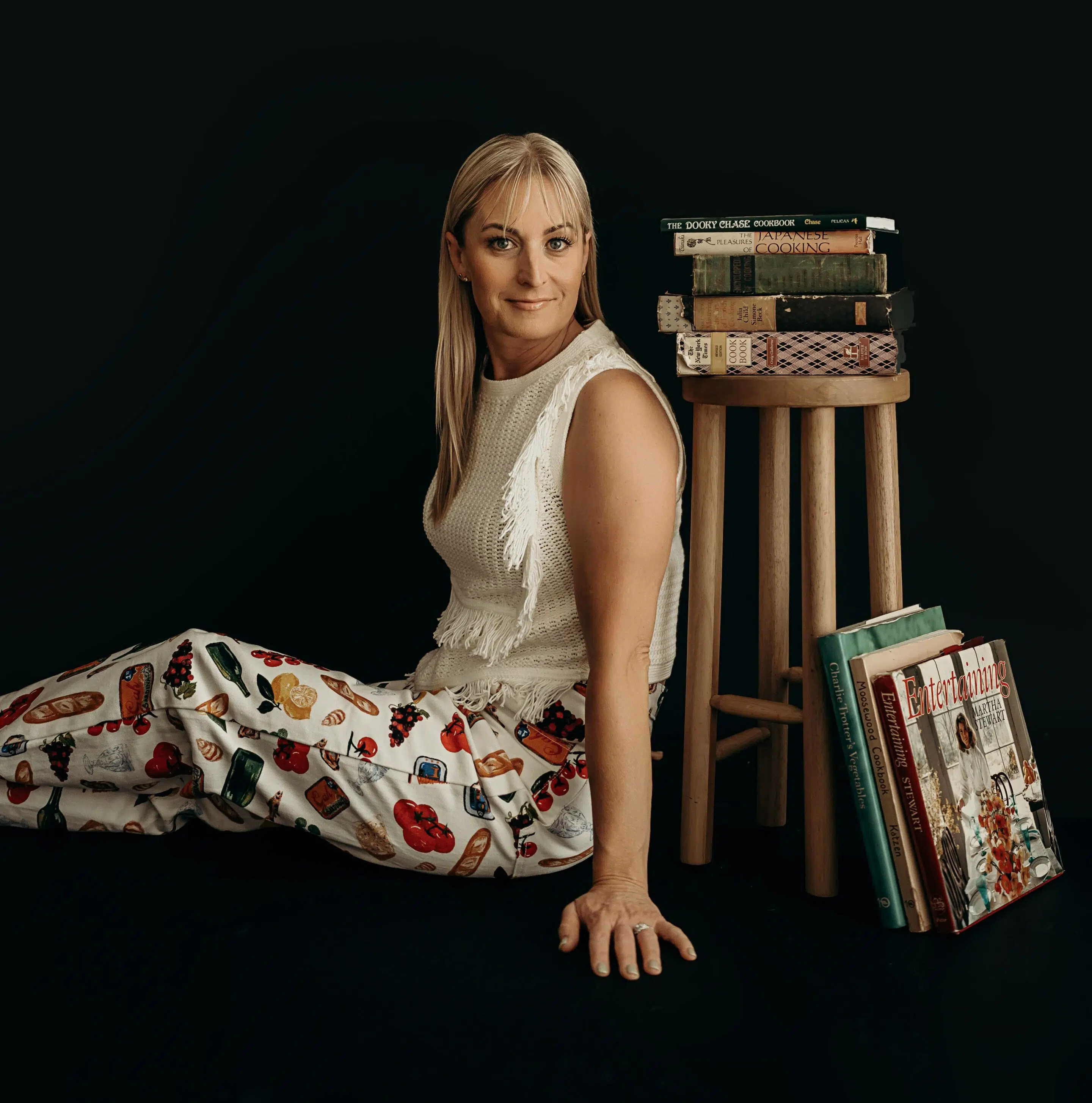 Chef Deb leans against a stool with an assortment of vintage cookbooks